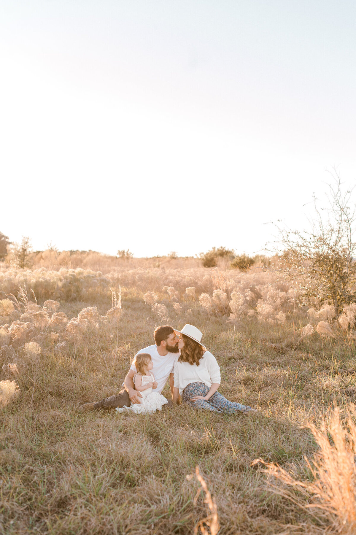 Family sits in a field at sunset while mom and dad kiss and mom holds her pregnant belly.