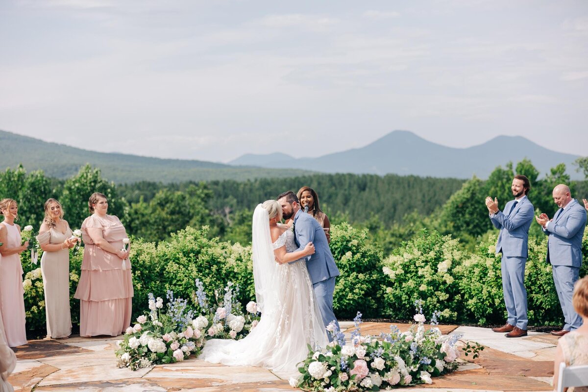 A bride and groom share a kiss at an outdoor wedding ceremony while guests look on and applaud.