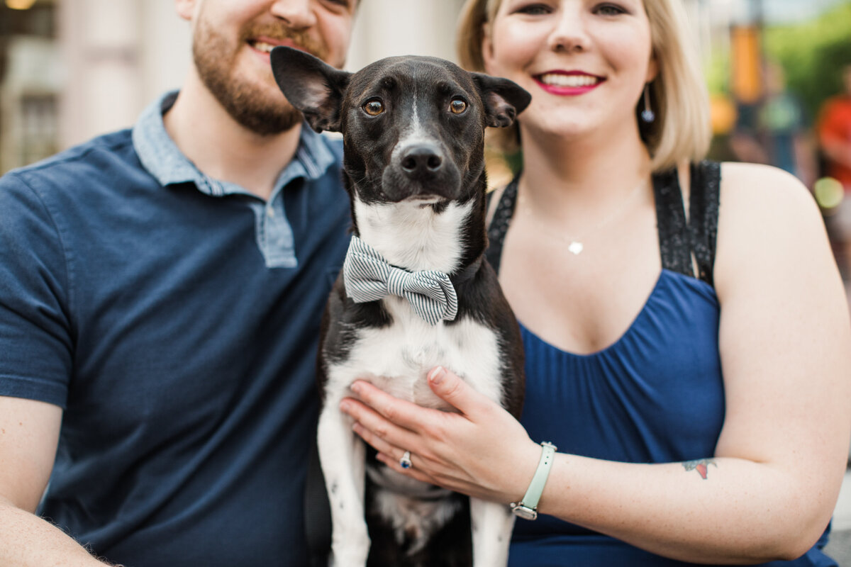 A photo of a couple where the main focus is on their dog during their engagement session in Fort Worth, Texas. The woman on the right is wearing a sleeveless blue top while the man on the left is wearing a short sleeve collared blue shirt. Their black and white is in the  middle and is wearing a striped white and blue bowtie.
