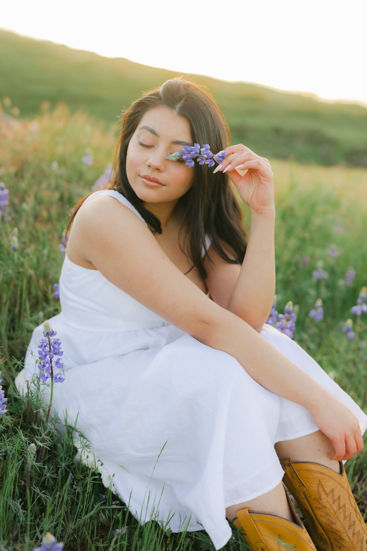 A dreamy editorial portrait of a woman sitting in a wildflower field at sunset in Greenville, SC. She wears a white dress and brown cowgirl boots, holding a purple lupine to her eye. Photographed by Dani Lacey.