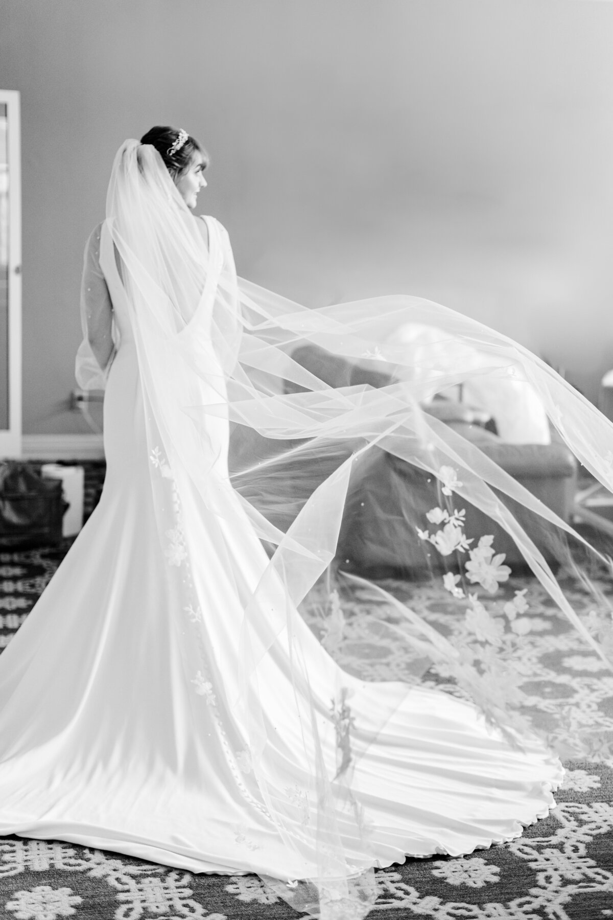 Black and white photo of bride glancing out the window with her veil flying behind her