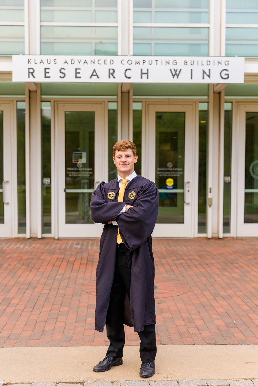 Graduate boy standing on front of Georgia tech Research wing on campus wearing graduation gown arms crossed during senior photos with Atlanta photographer Laure Photography 
