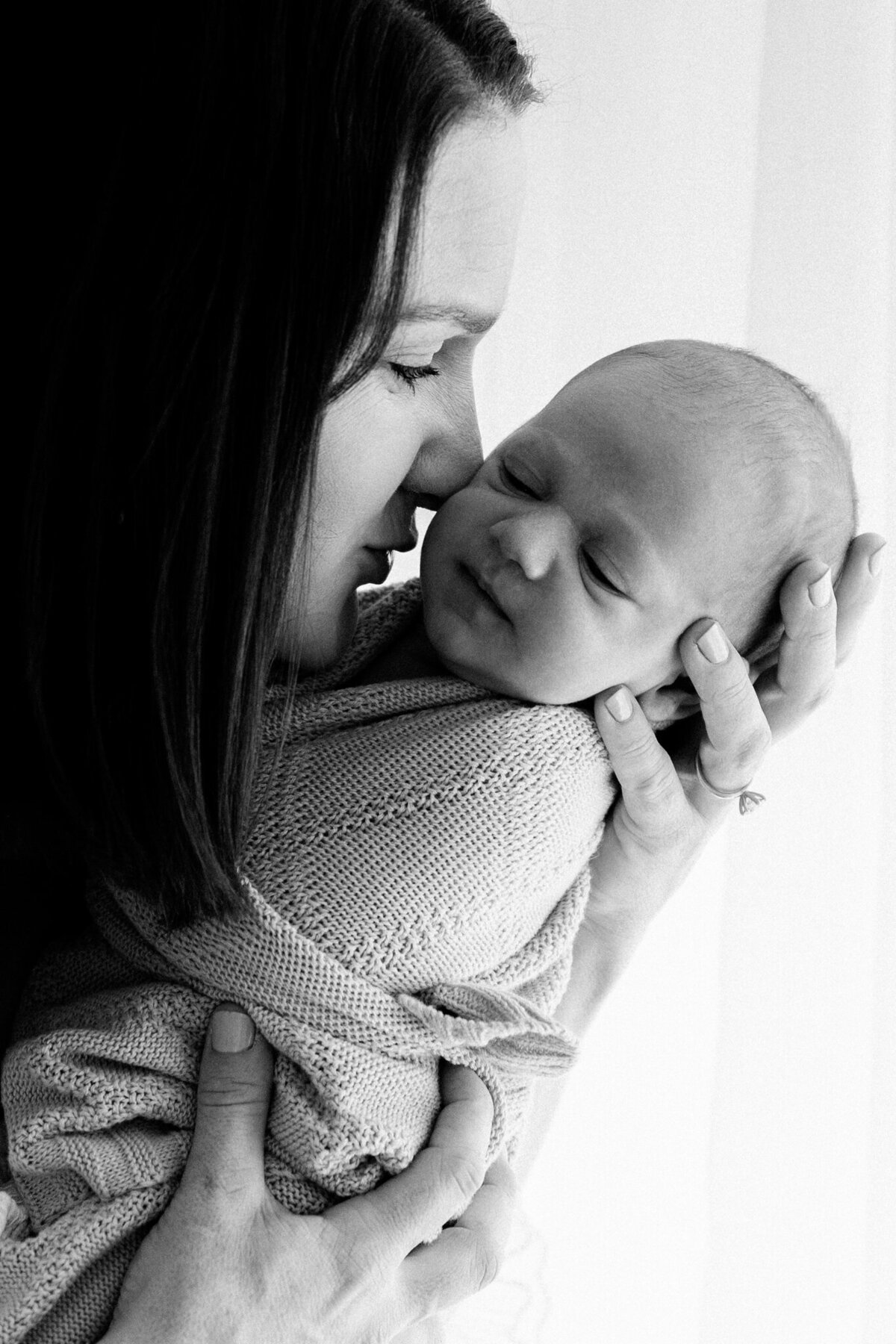 Black and white portrait of a mother holding her swaddled newborn close, capturing a tender moment during a studio newborn session in Southwest Sydney.