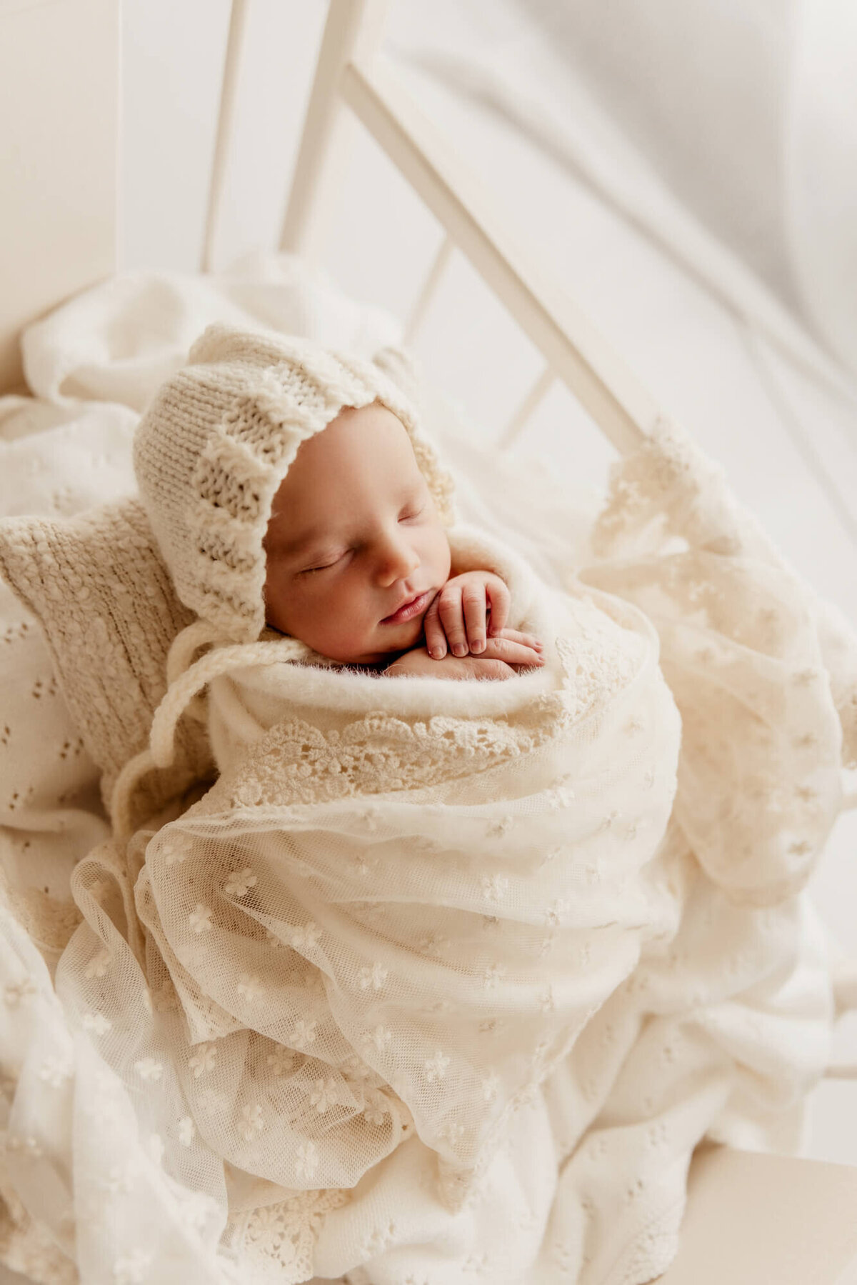 Newborn baby girl wrapped in a white swaddle with her hands near her chin sleeping peacefully.