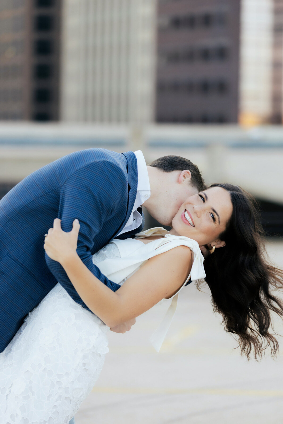 Couple in Dallas downtown during their engagement session, he dips her for a neck kiss as her hair flies and she laughs at the camera.