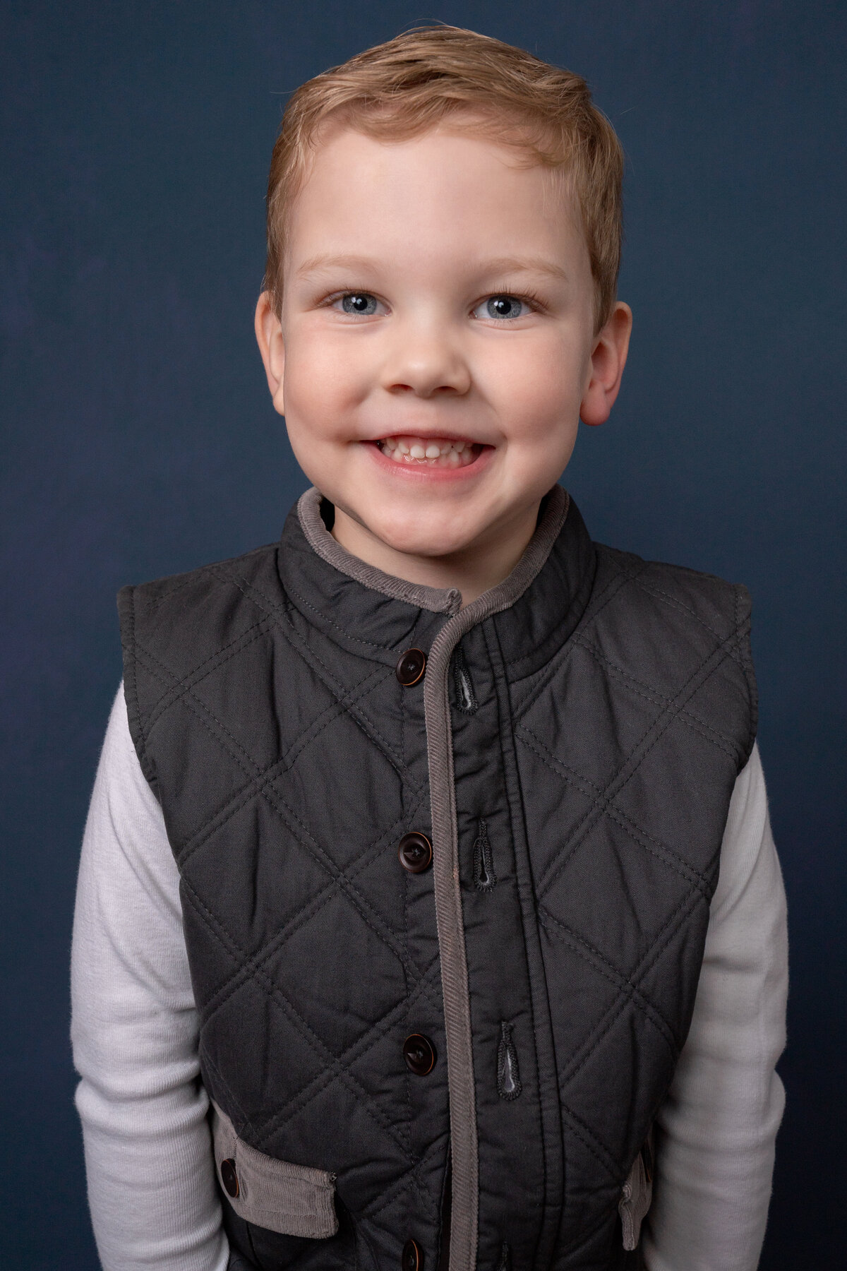 Closeup portrait of young boy wearing quilted vest and white shirt in front of blue backdrop. Child is smiling.