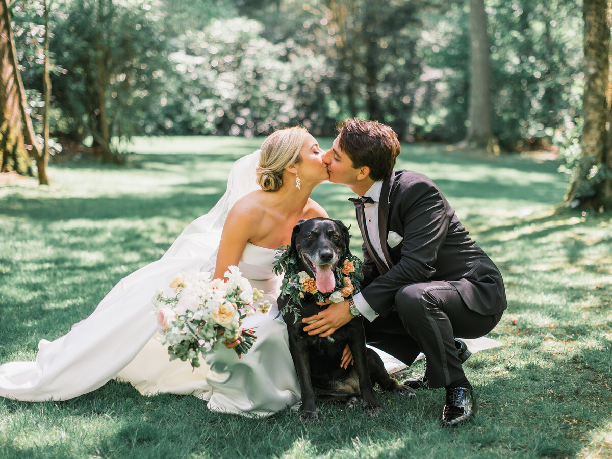Bride and groom share a kiss on the lawn with their black dog wearing a floral collar at Old Edwards Inn in Highlands, North Carolina.