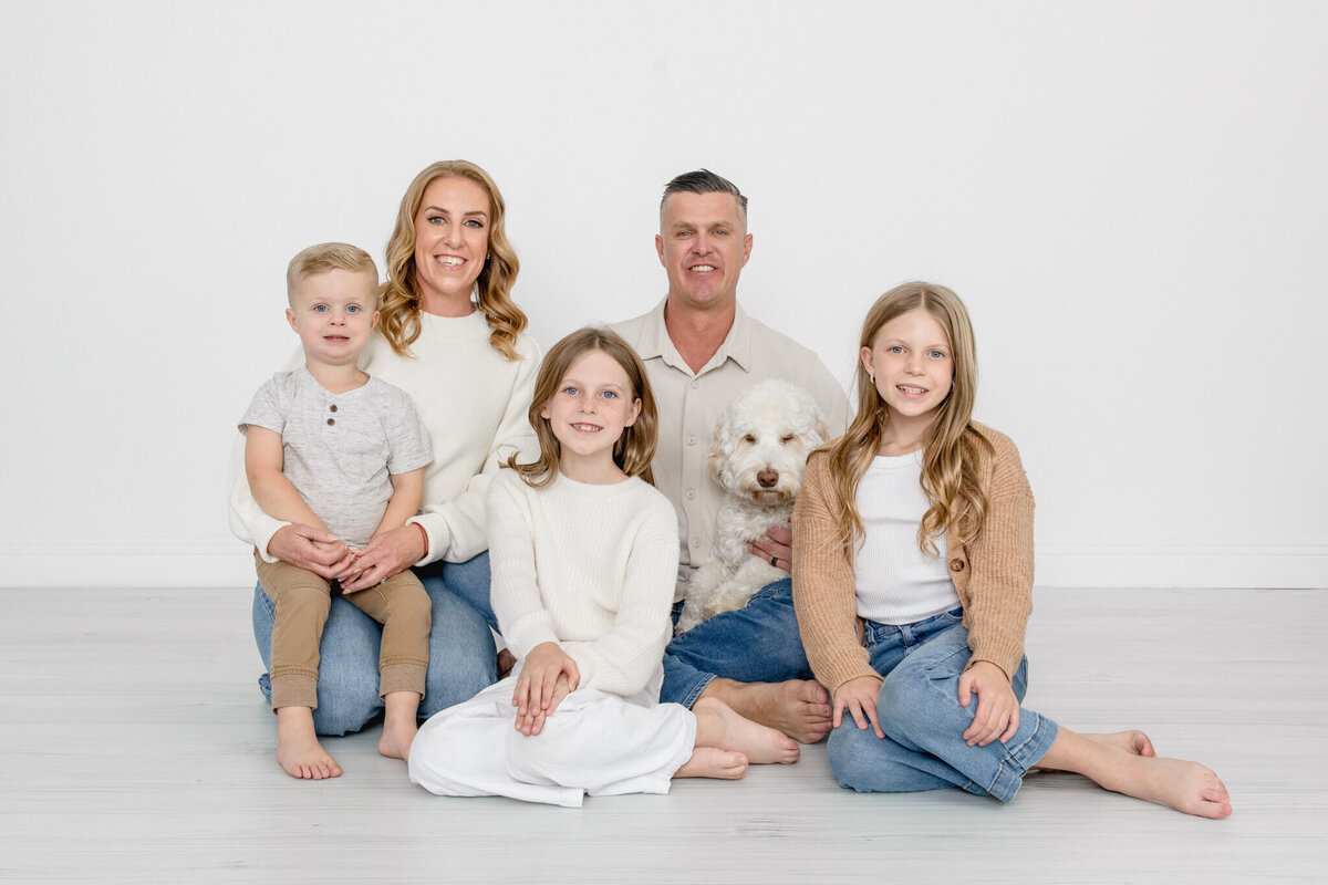 A family of five sitting together on a grey studio floor with their dog, all with blonde hair and wearing shades of white, brown, and blue, smiling at the camera against a white background.
