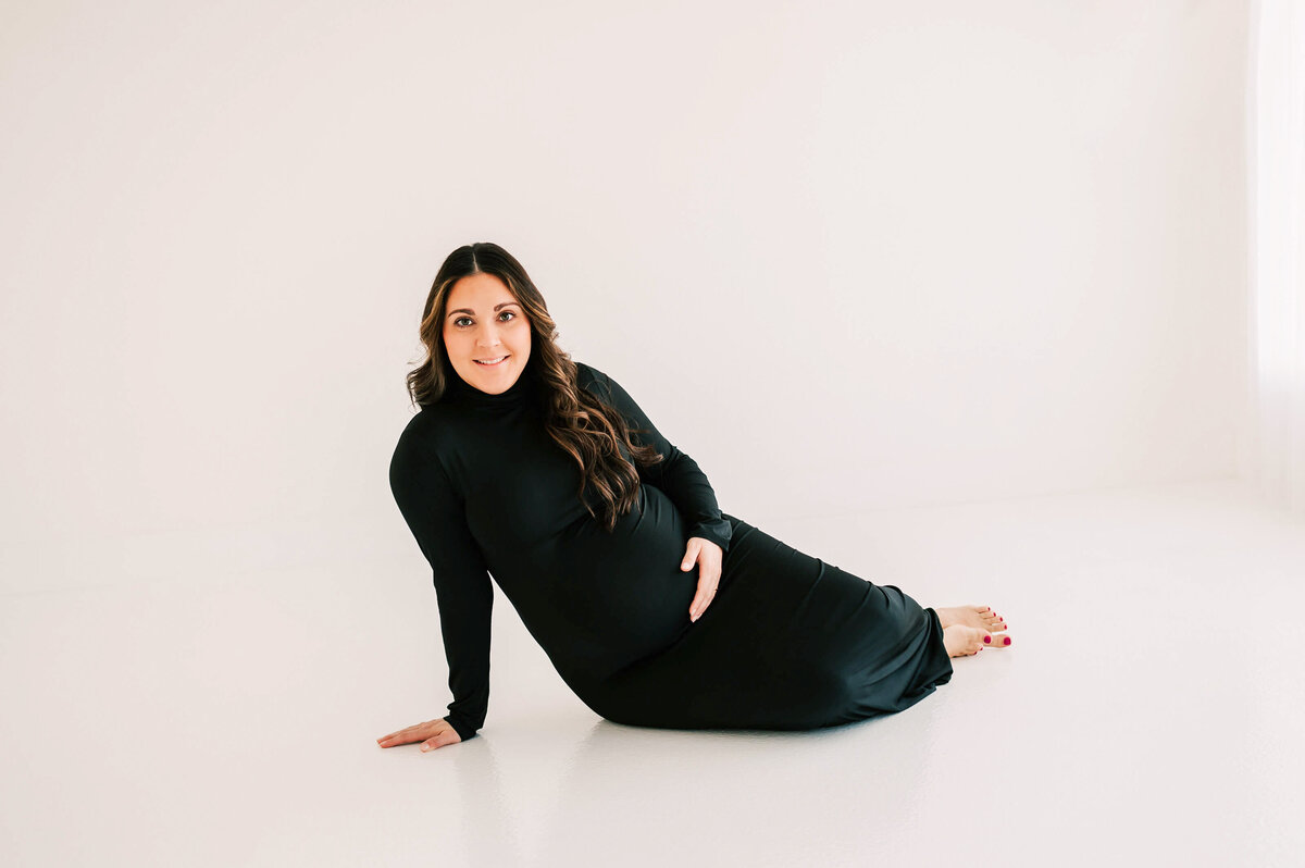 pregnant mom in black dress smiling while sitting on the floor of studio
