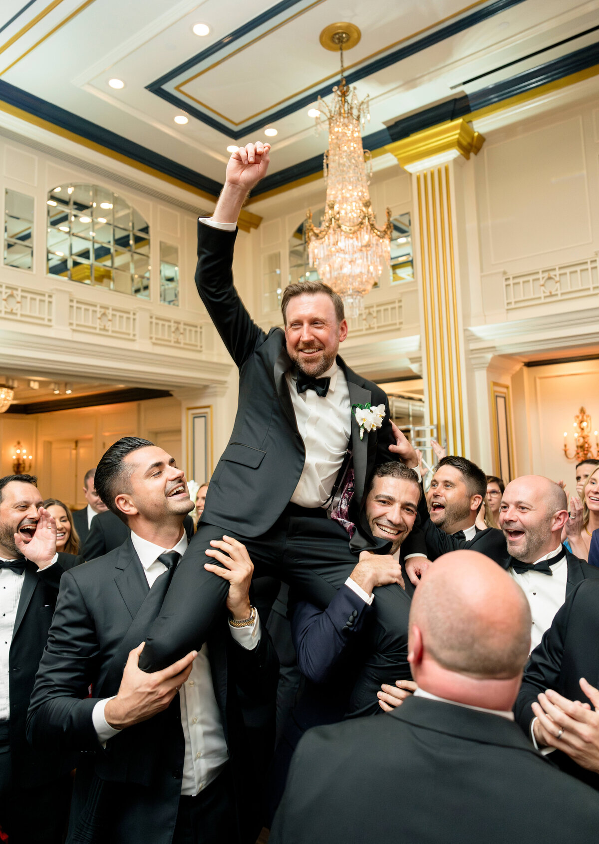 A joyful man in a tuxedo is lifted by a group of smiling, well-dressed people in an elegant ballroom with chandeliers. The celebratory atmosphere is enhanced by their happy expressions and the luxurious setting.
