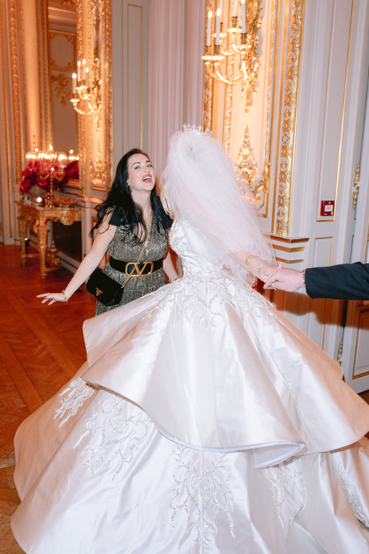 The enchanting moment captured in the photo was taken at the opulent Shangri La Hotel & Ritz Paris Hotel in Paris, France, by the talented photographer Matteo Coltro. In the scene, a woman in a striking black dress, accentuated by a large belt, warmly reaches out towards a radiant bride adorned in a voluminous white gown with exquisite embroidery. The bride's ensemble is elegantly complemented by a delicate veil and sparkling tiara. The setting exudes luxury, with an ornate room filled with shimmering chandeliers and intricate gold detailing adorning the walls, providing a majestic backdrop for this touching interaction between the two women.   Additionally, the magic of this moment was beautifully captured on film by the talented videographers at Zen Film Works, enhancing the visual storytelling and capturing the emotions and beauty of this special occasion.