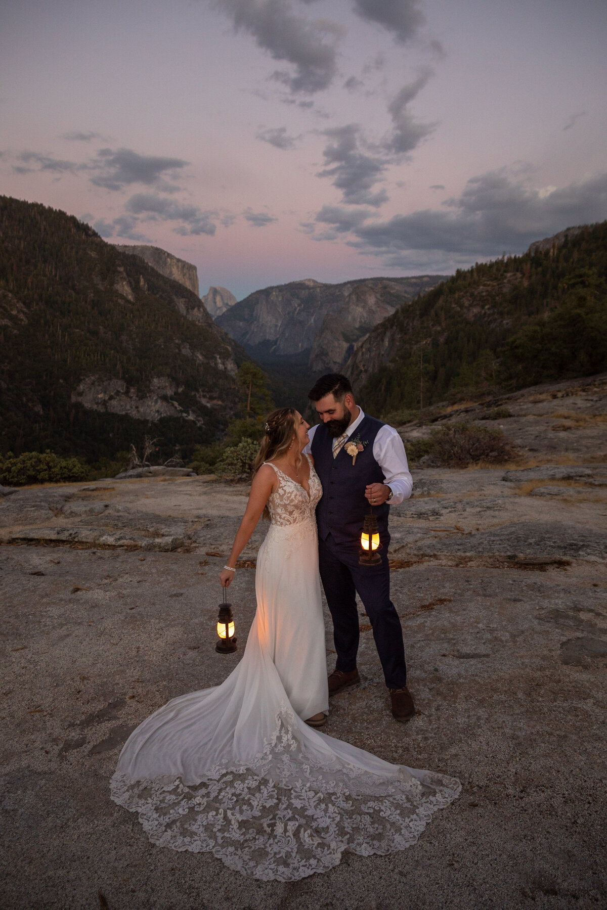 A bride and groom stand next to each other holding lanterns by their side as the sunsets over Half Dome behind them.
