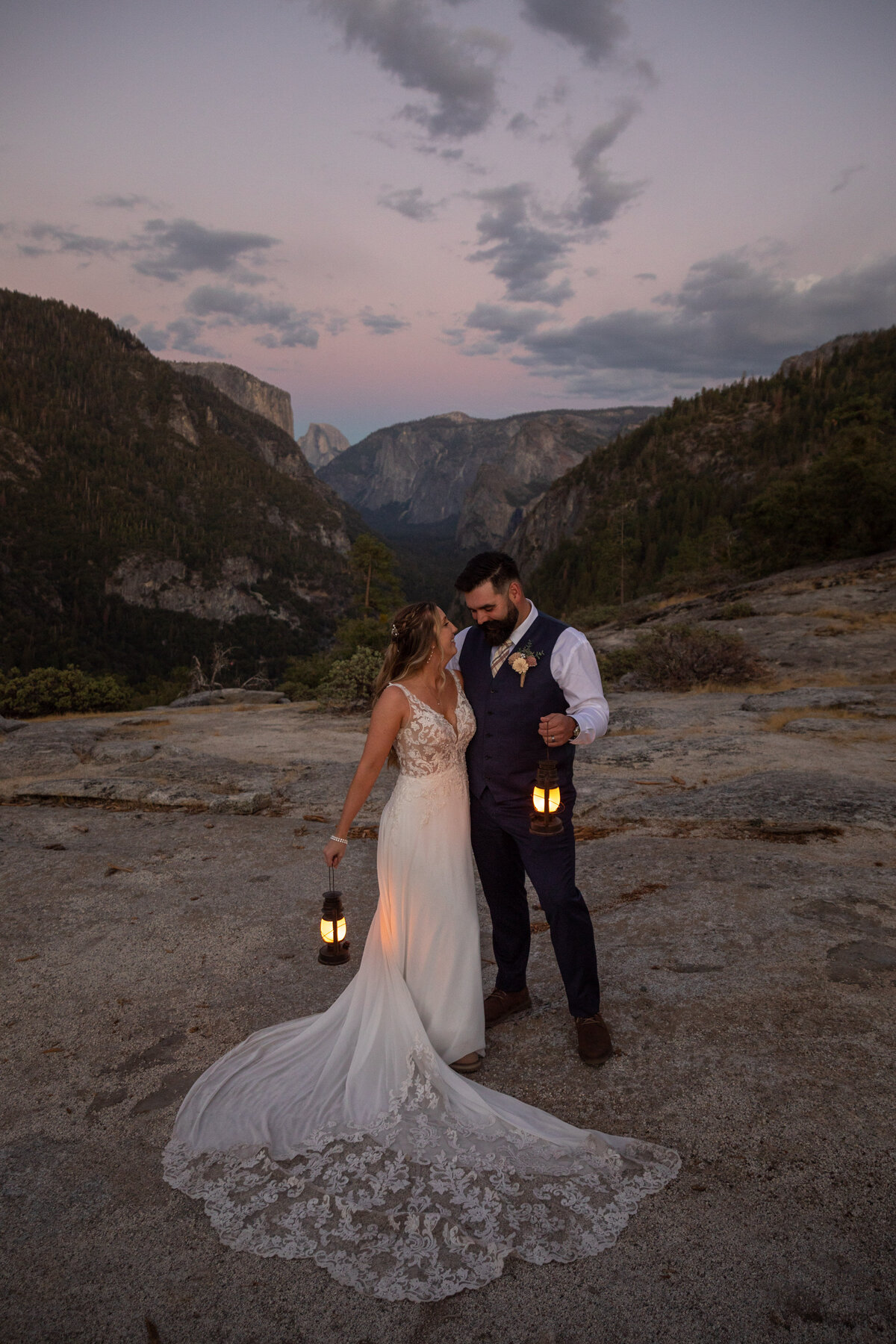 A bride and groom stand next to each other holding lanterns by their side as the sunsets over Half Dome behind them.