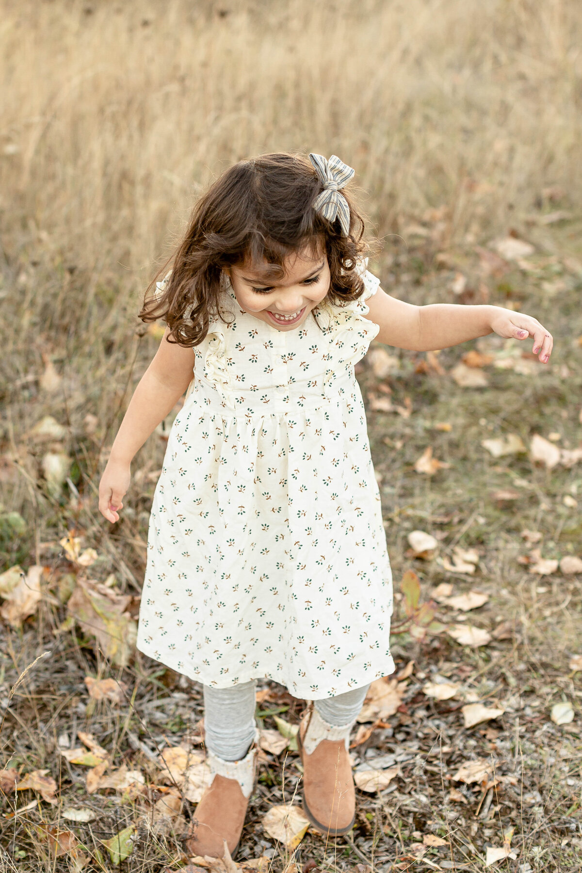 Toddler girl with cream dress on standing in a field and smiling looking down at the ground during fall family photography session.