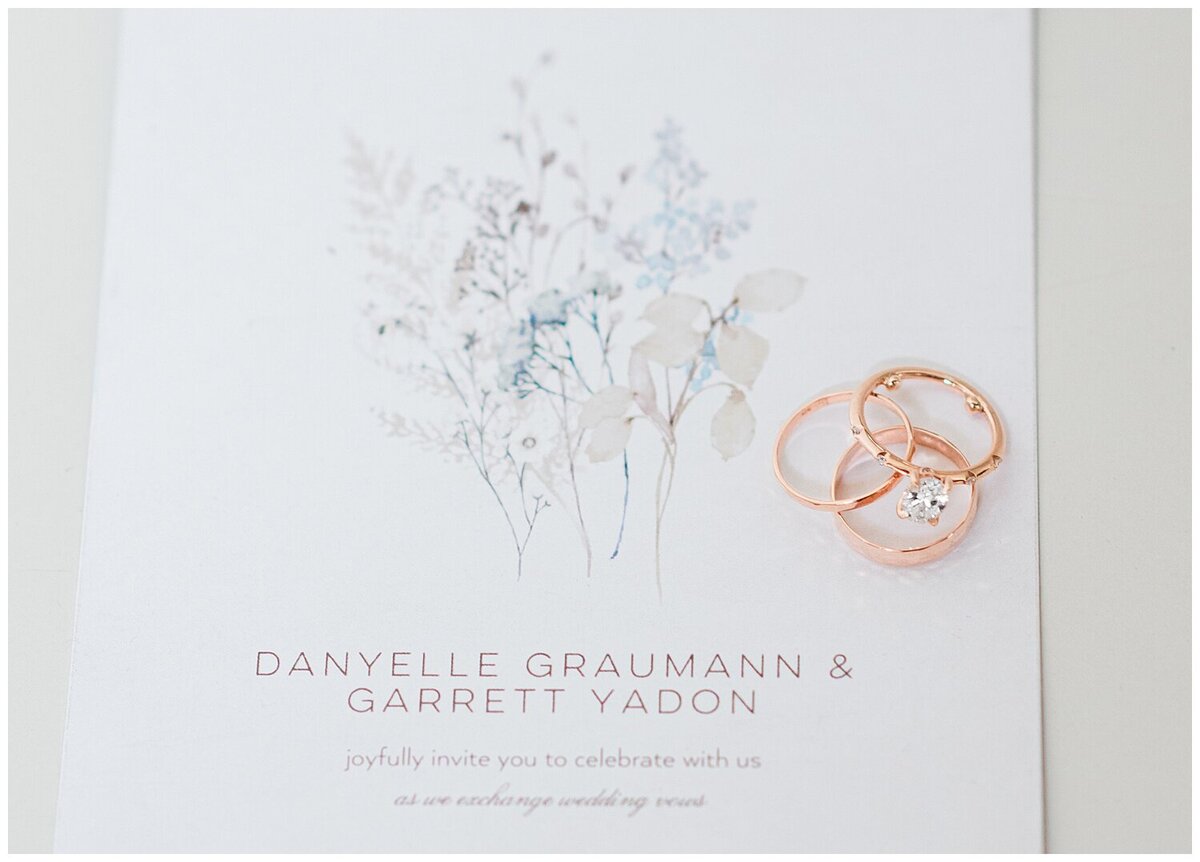 Close up photo of a white wedding invitation  on a white table with the wedding rings on the right side of the invitaion