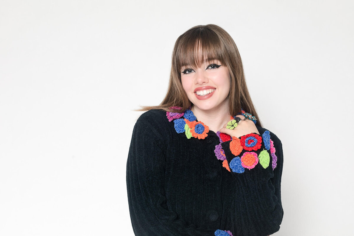 A person with long straight hair wearing a black sweater adorned with colorful crocheted flowers smiles against a plain white background.