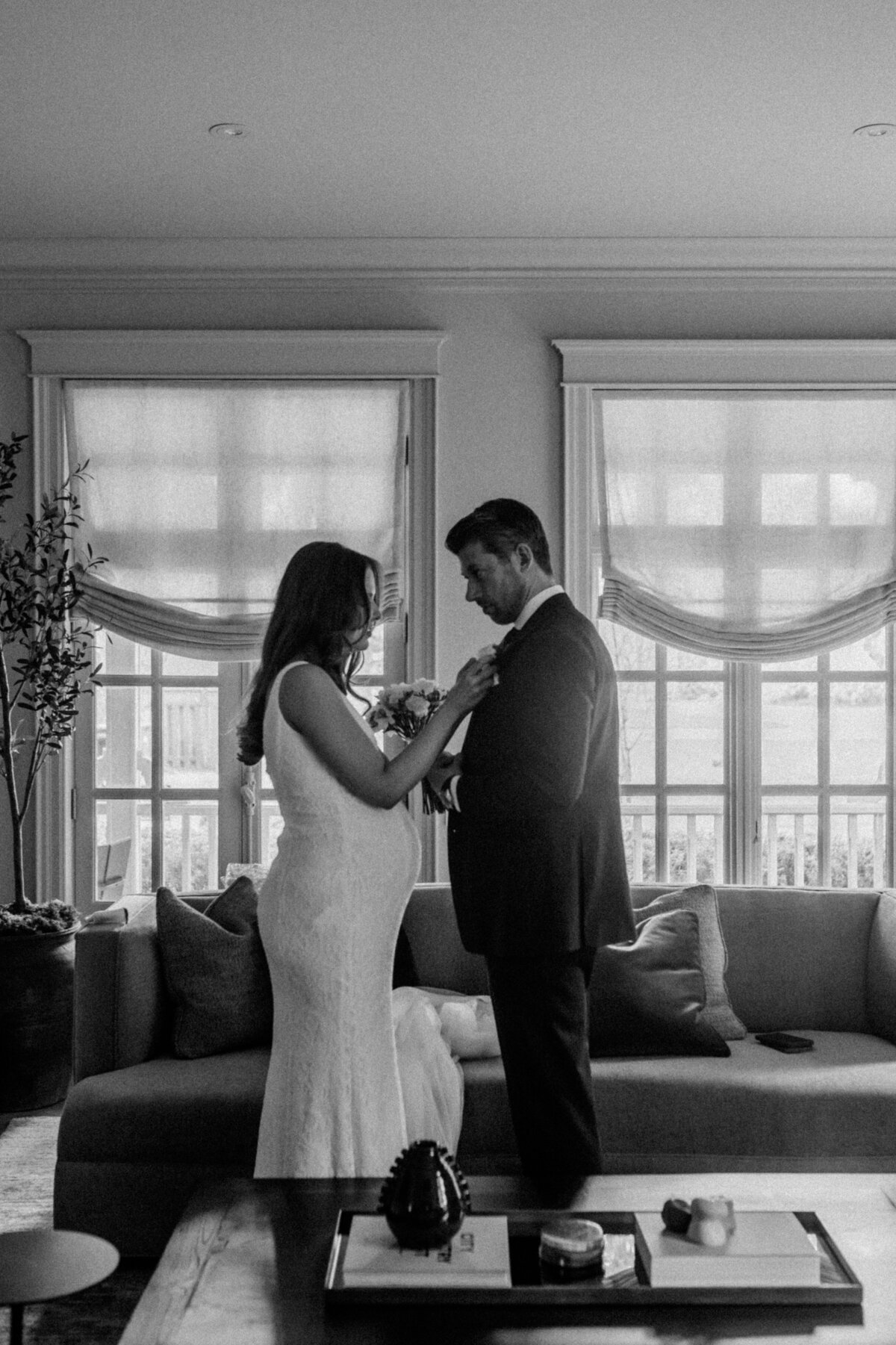 A heartfelt black-and-white photograph of a pregnant bride adjusting her groom's boutonniere, sharing a quiet moment together.