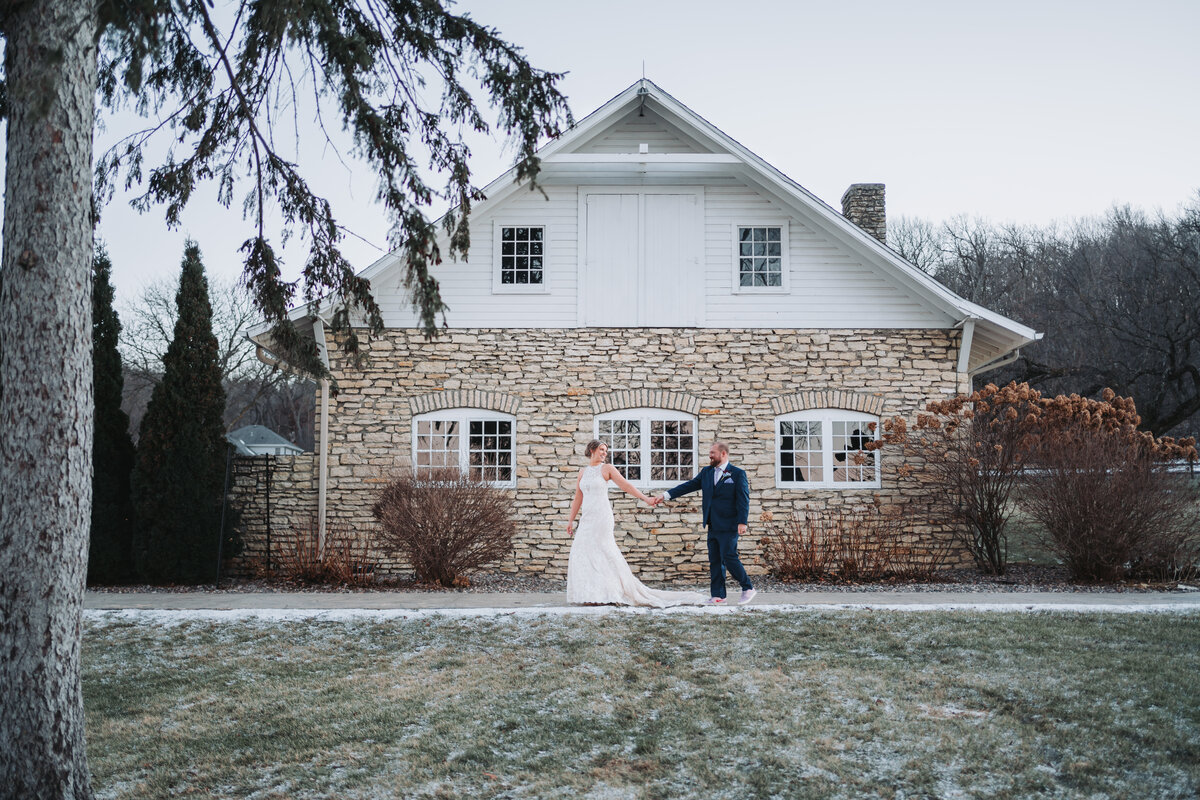 A couple stands outside a rustic stone and white farmhouse on a winter day.