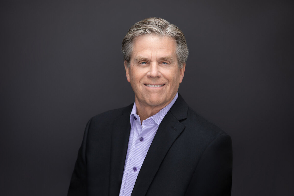 A businessman with grey hair and purple shirt in a black suit poses for a professional headshot on a black backdrop for Janel Lee Photography studios in Cincinnati Ohio