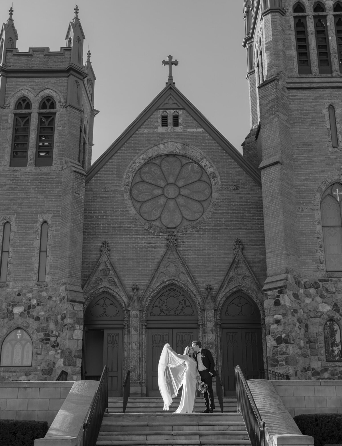 bride and groom embrace outside ornate historic cathedral, St. Paul on the Lake, the bride's long lace veil like angel wings