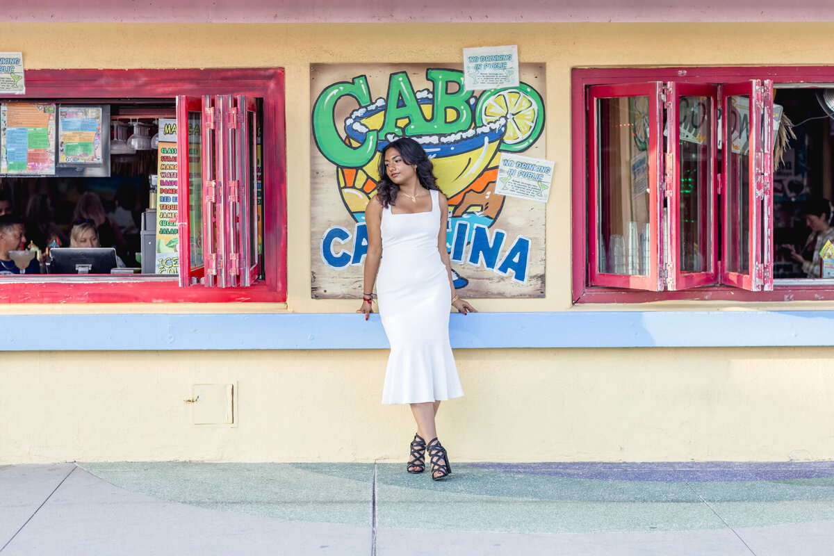 A woman in a white dress stands outside Cabo Cantina, leaning against the buildings yellow wall. The cantina has red-framed open windows, colorful signs, and a lively interior visible in the background.