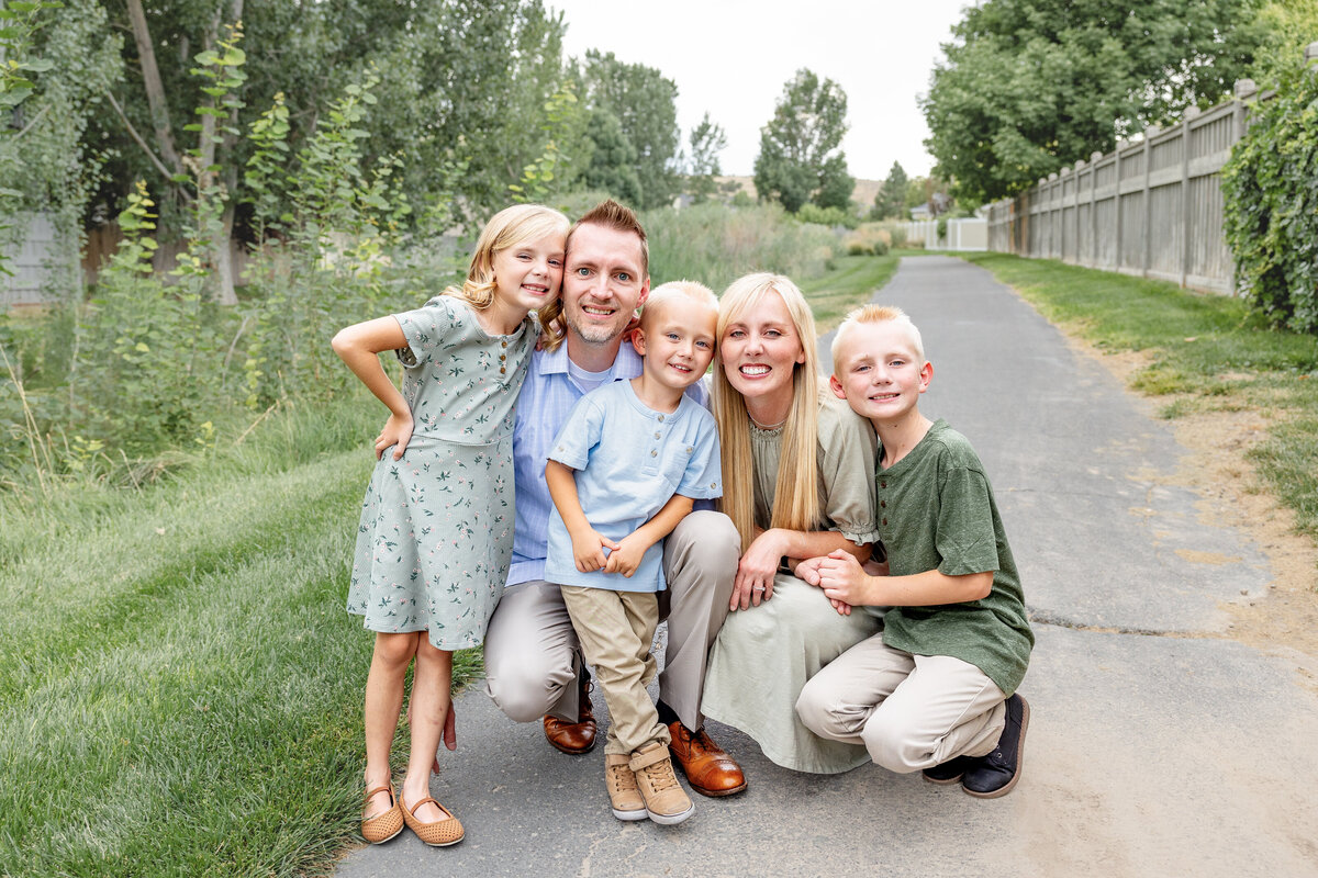 A happy family of five crouching together on a tree-lined pathway, smiling at the camera. Captured by Cassie Lee Photography.