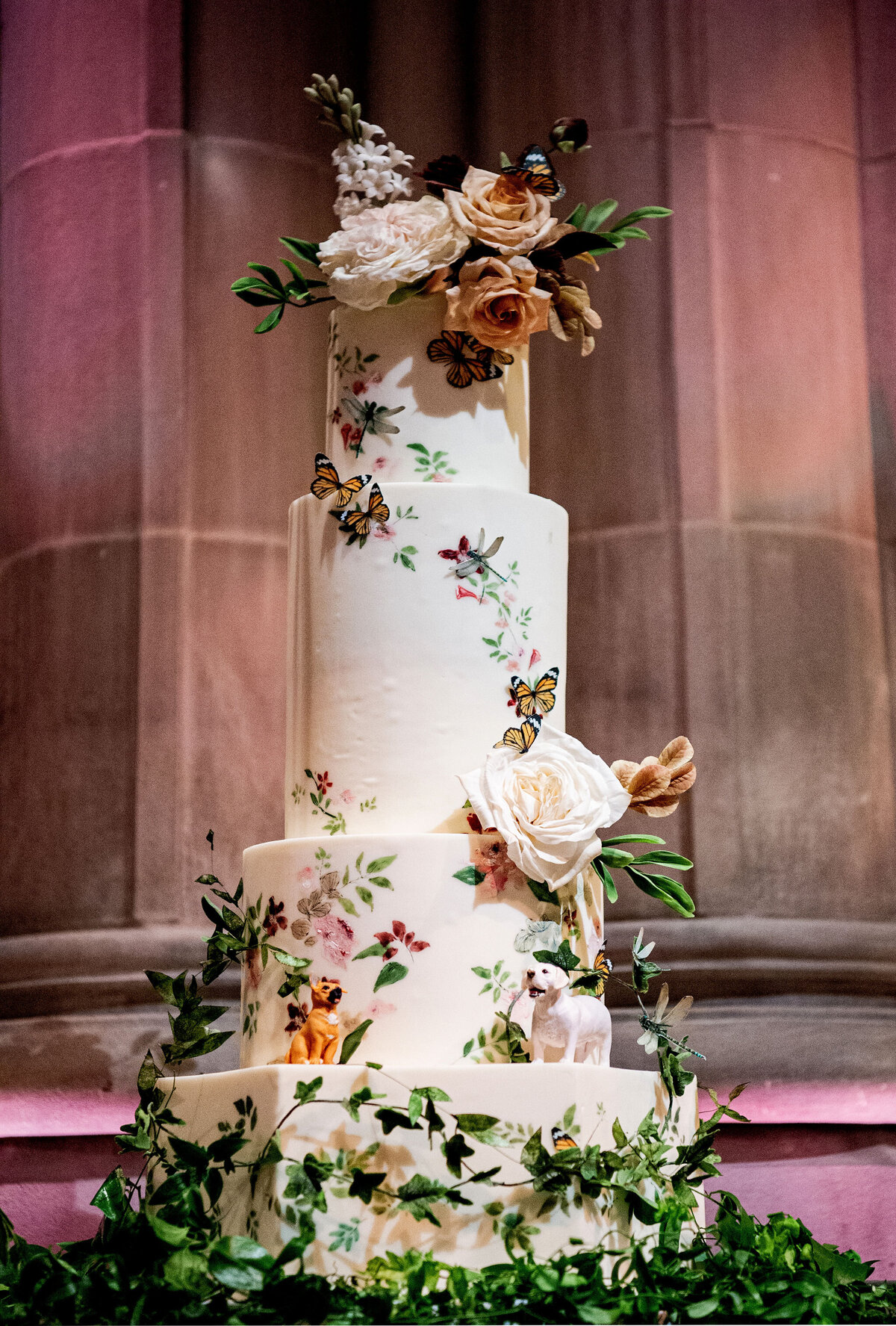 This photo was taken by photographer Twah Dougherty at Washington National Cathedral in Washington D.C. The wedding cake featured in the photo was created by Current Films as Videographers. The tall cake has four tiers and is decorated with flowers, butterflies, and greenery, with intricate floral designs on each tier. Leafy green foliage surrounds the base, and there is a dog figurine placed on one of the tiers.