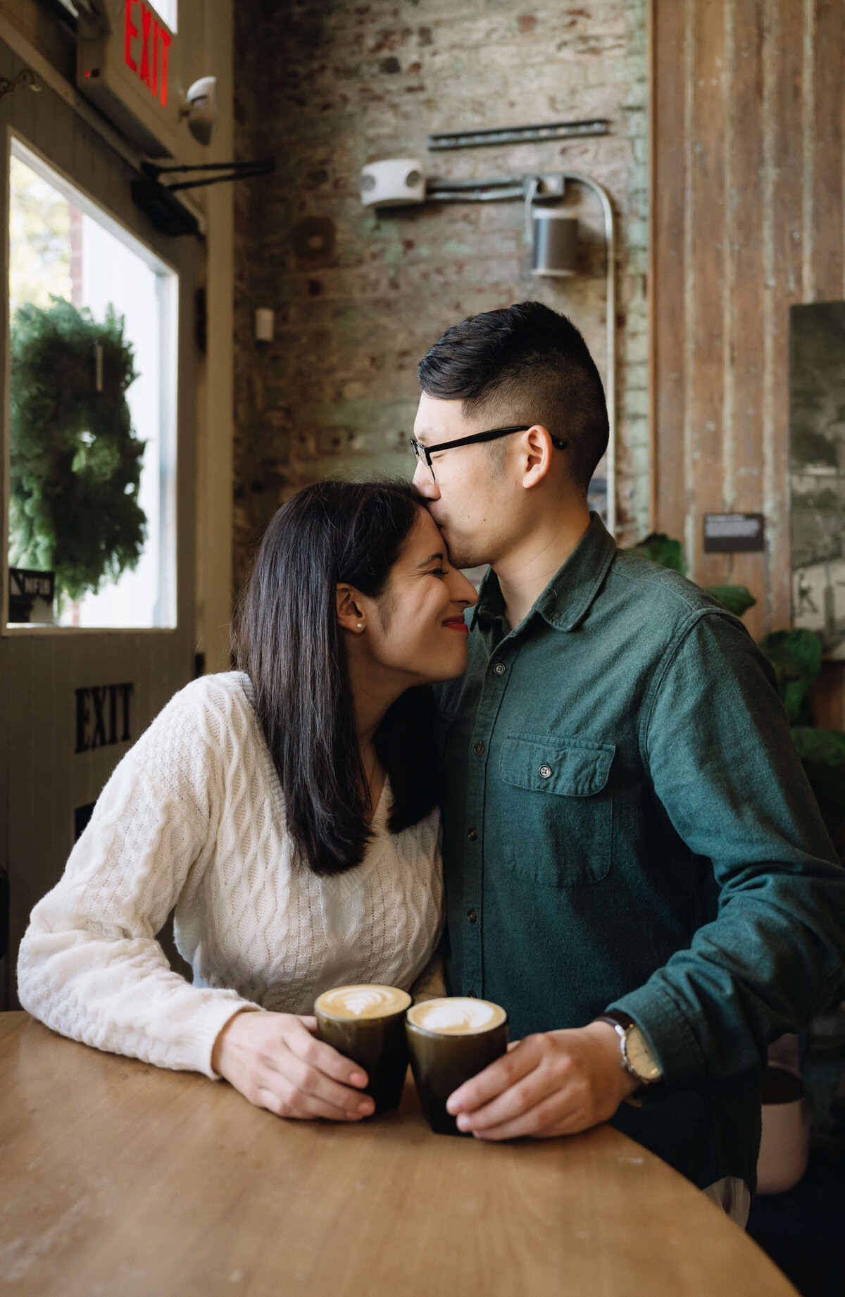 fall engagement photos indoors at a coffee shop with man kissing womans head as she smiles while they hold coffee together