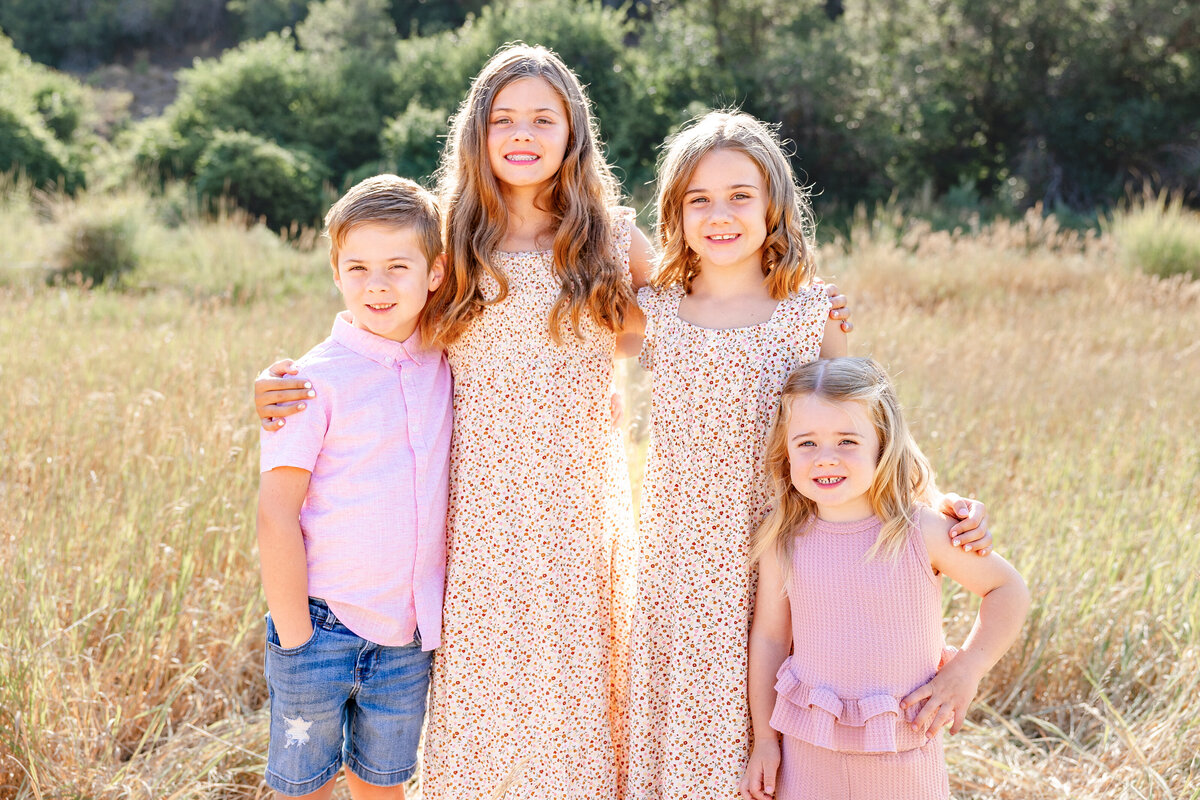 A group of children, dressed in floral and pastel-colored outfits, standing together and smiling in an open field.