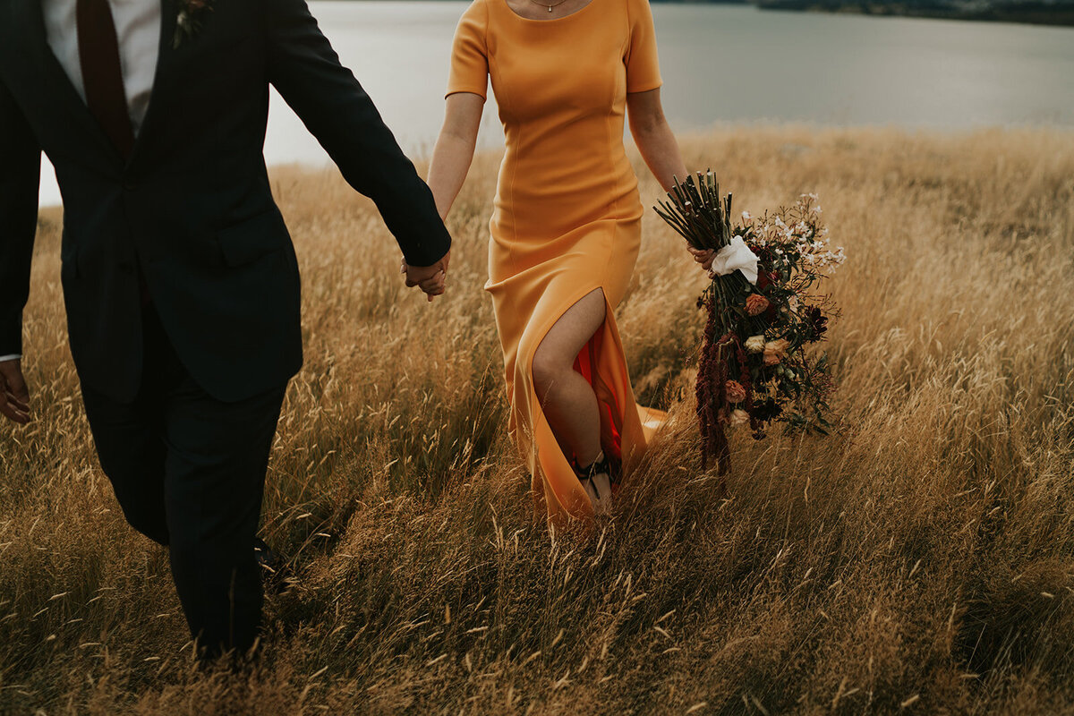 The Vase Floral Co - bride and groom walk through field, bride carries beautiful rustic coloured bouquet