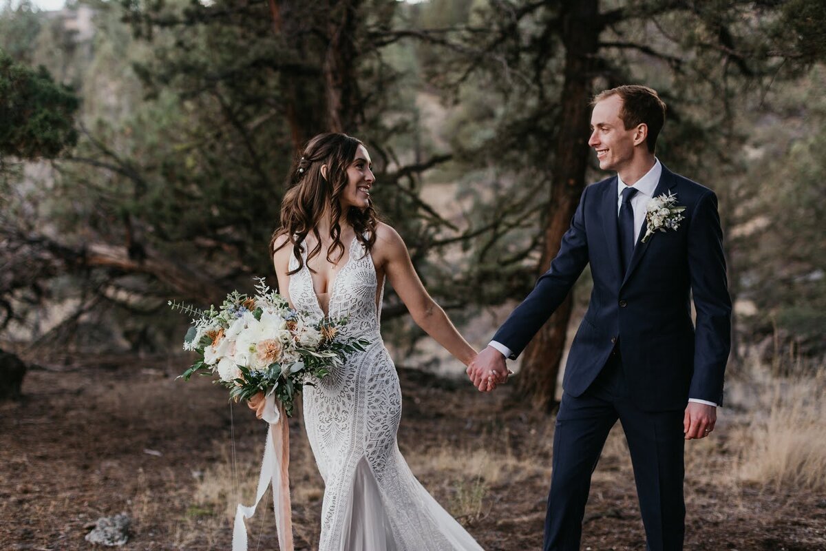 Bride and groom hold hands and look at each while she is holding a lush floral bouquet.