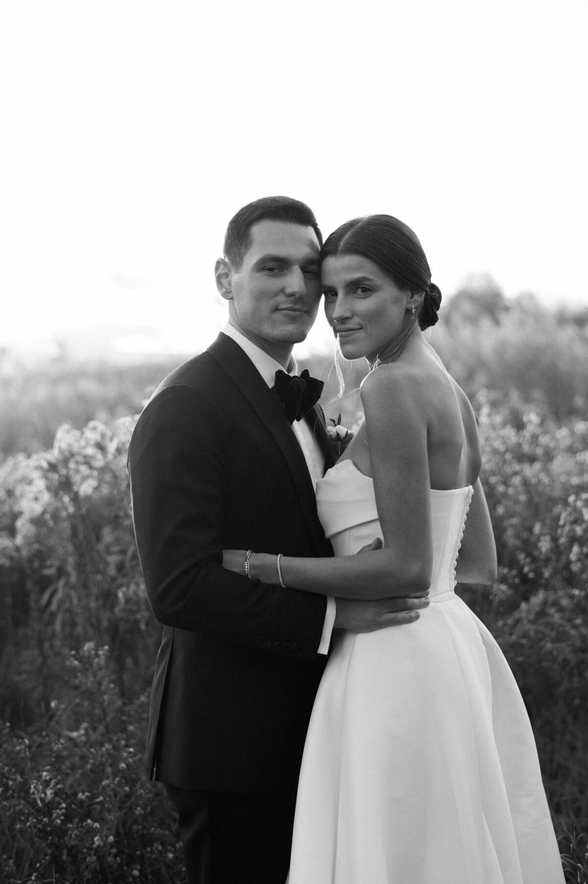 The bride and groom stand closely in a field, gazing into the camera with a serene expression