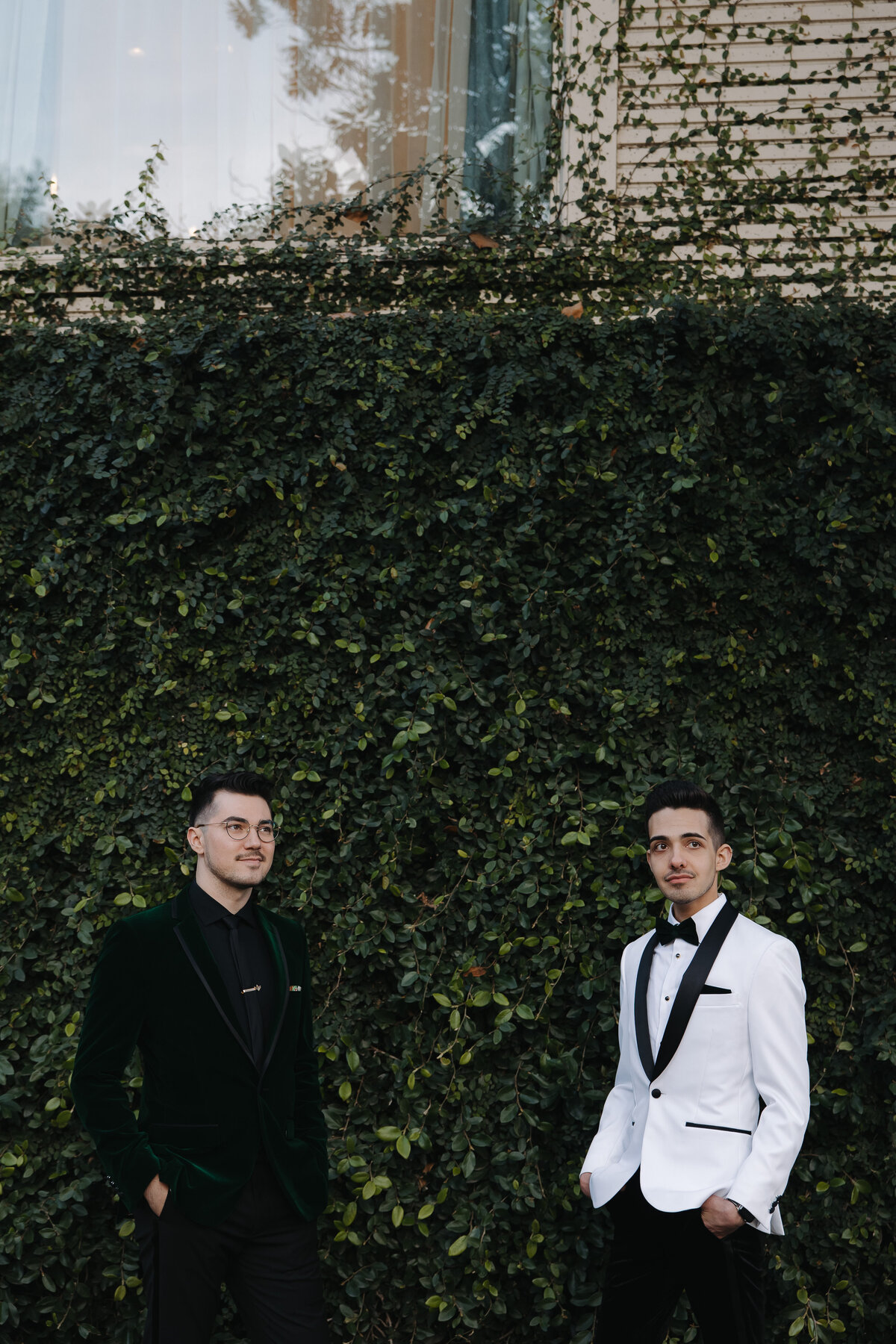 Two stylish grooms pose against an ivy-covered wall, showcasing artistic and editorial wedding photography with a refined, luxury aesthetic.