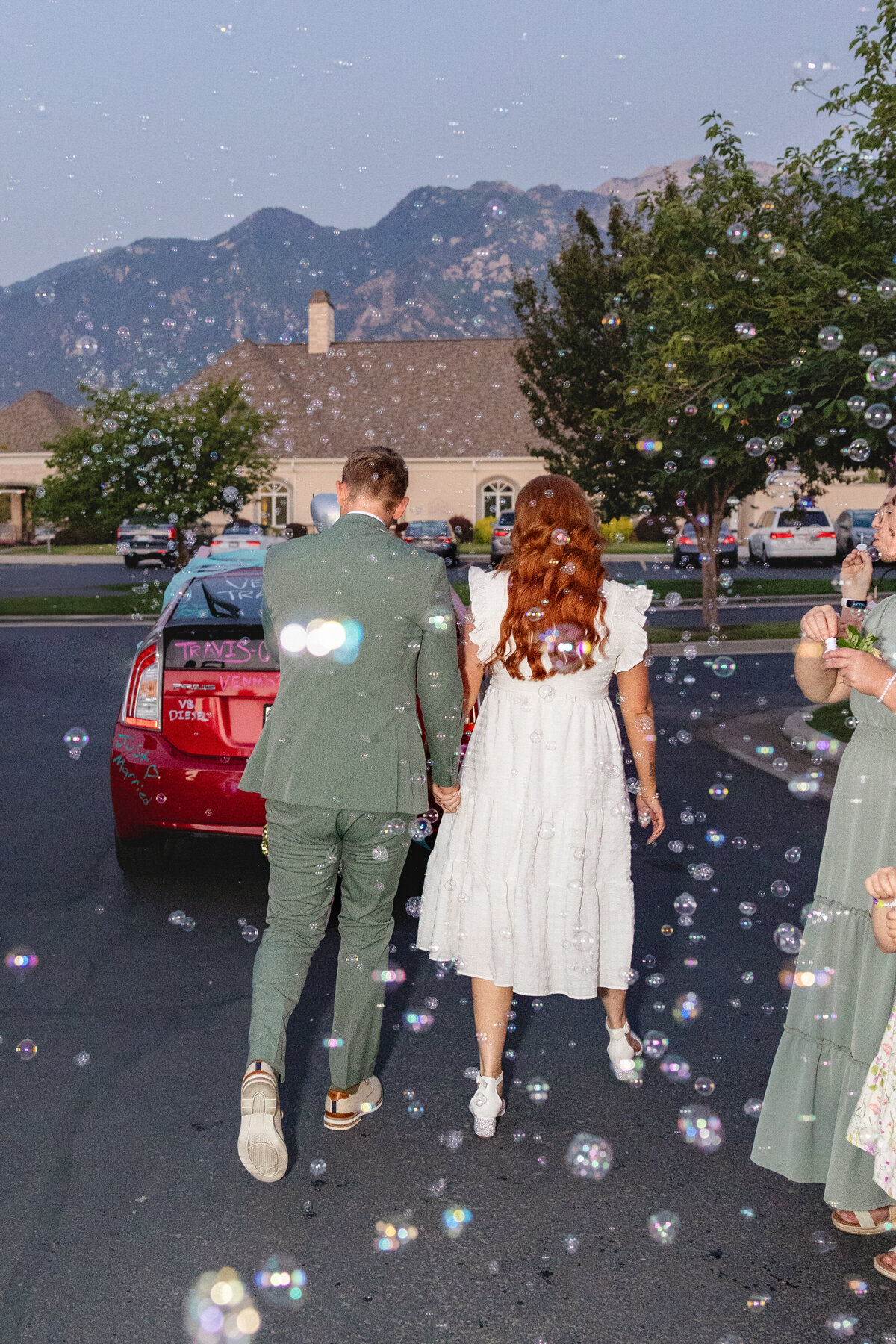 The bride and groom walk away hand in hand towards their decorated getaway car, with bubbles floating around them and the beautiful Utah mountains in the background.