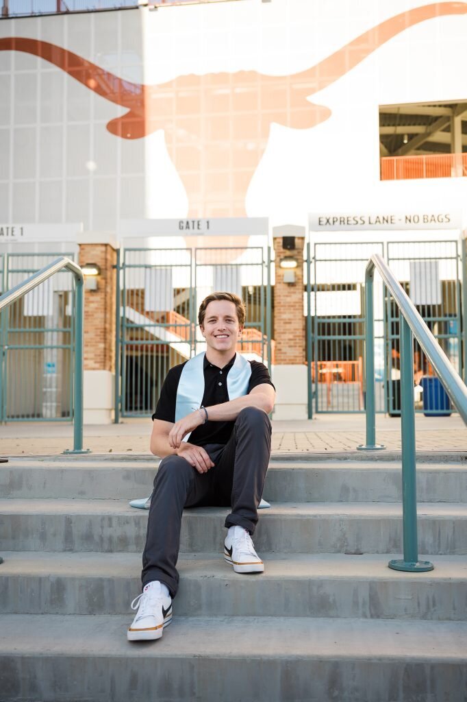 Young man sitting on stairs for graduation photos outside a stadium entrance, wearing a light blue stole. Background features a large University of Texas Longhorn logo in orange.