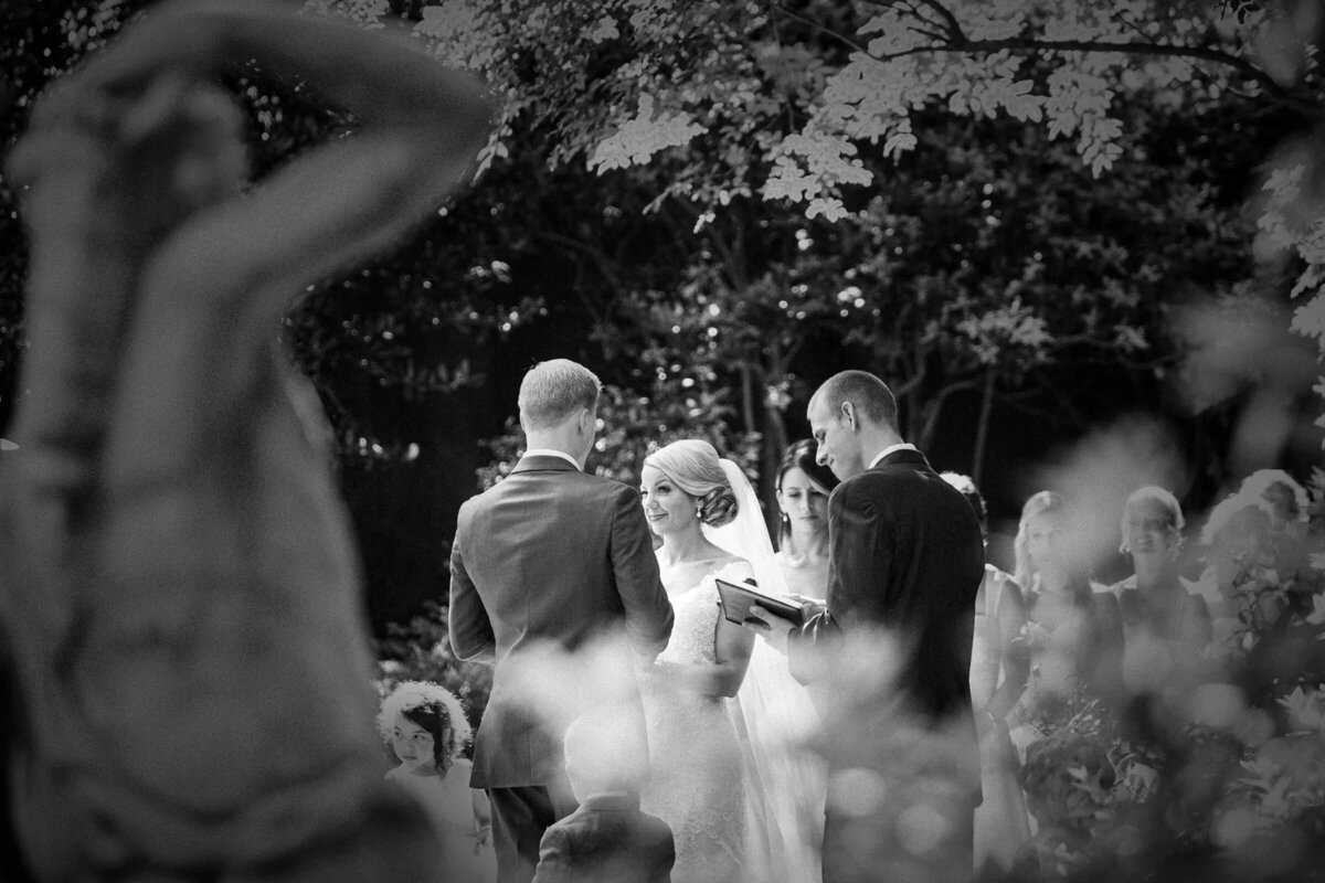 Bride and groom exchanging vows in an outdoor setting, partially viewed through foliage