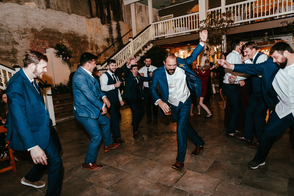 A group of men in suits joyfully dance together at an outdoor event, with one man in the center raising his arms, surrounded by others clapping and cheering. The setting is lively, festive, and warmly lit.
