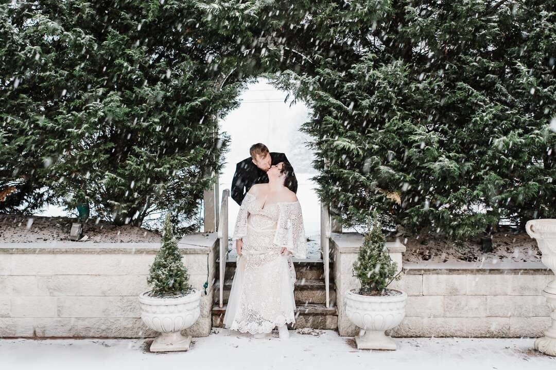 Bride and groom share a romantic kiss in the snow during a winter wedding, framed by evergreen trees and stone steps at an elegant New Jersey venue.
