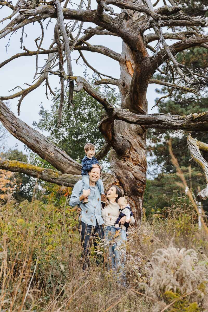 In an authentic family portrait set in Toronto, a family of four stands before a towering bare tree. One child joyfully sits on the man's shoulders, while the woman lovingly carries the other. They are enveloped by tall grass and lush foliage, capturing a genuine moment of togetherness.