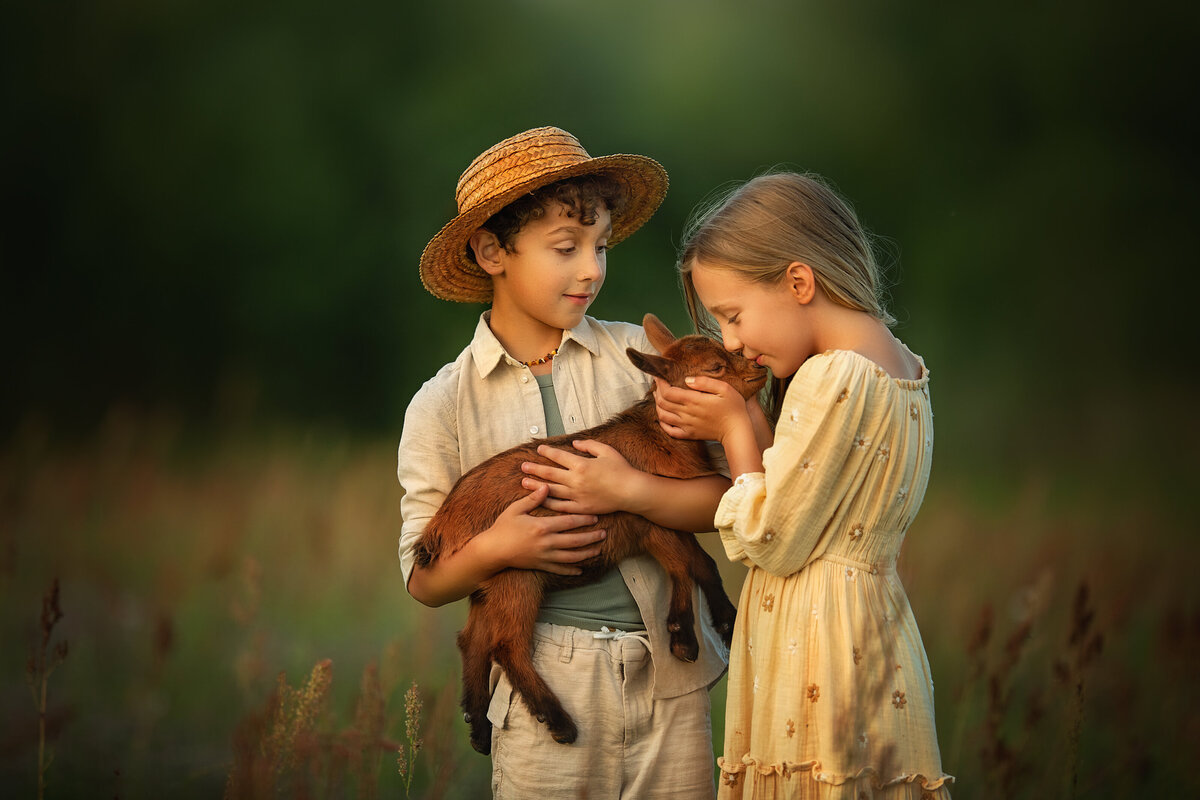 farmer boy with linen clothes and straw hat is holding a baby goat while a girl in yellow dress is kissing it in a tall grass field in Lithuania.
