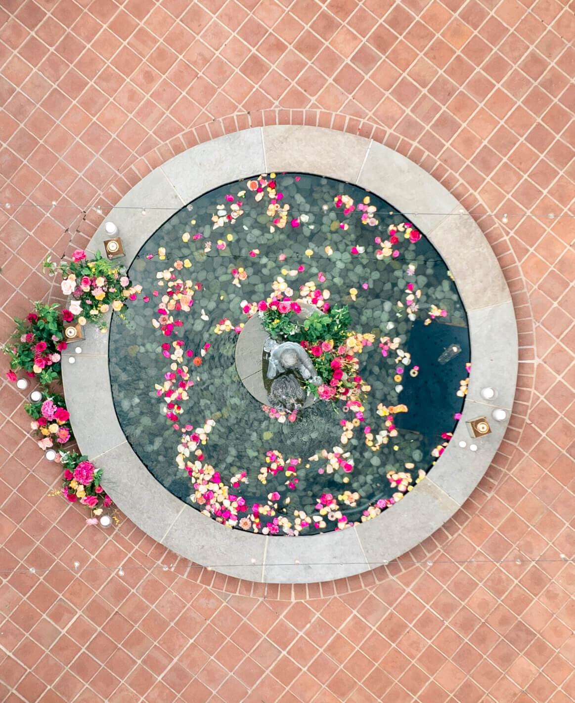 Aerial view of a circular water fountain with floating pink, yellow, and white flowers. The fountain is surrounded by a tiled, terracotta-colored floor with small decorative plants and candles on the perimeter.