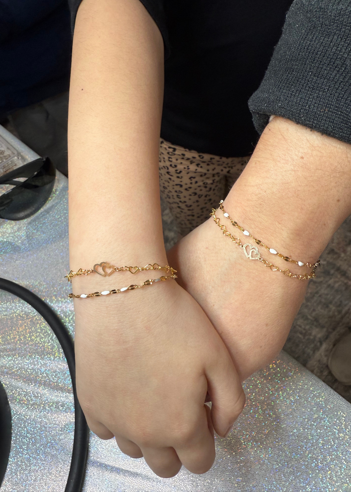 Matching gold heart chain bracelets on two wrists, symbolizing a sweet connection, styled with delicate mother-of-pearl accents.