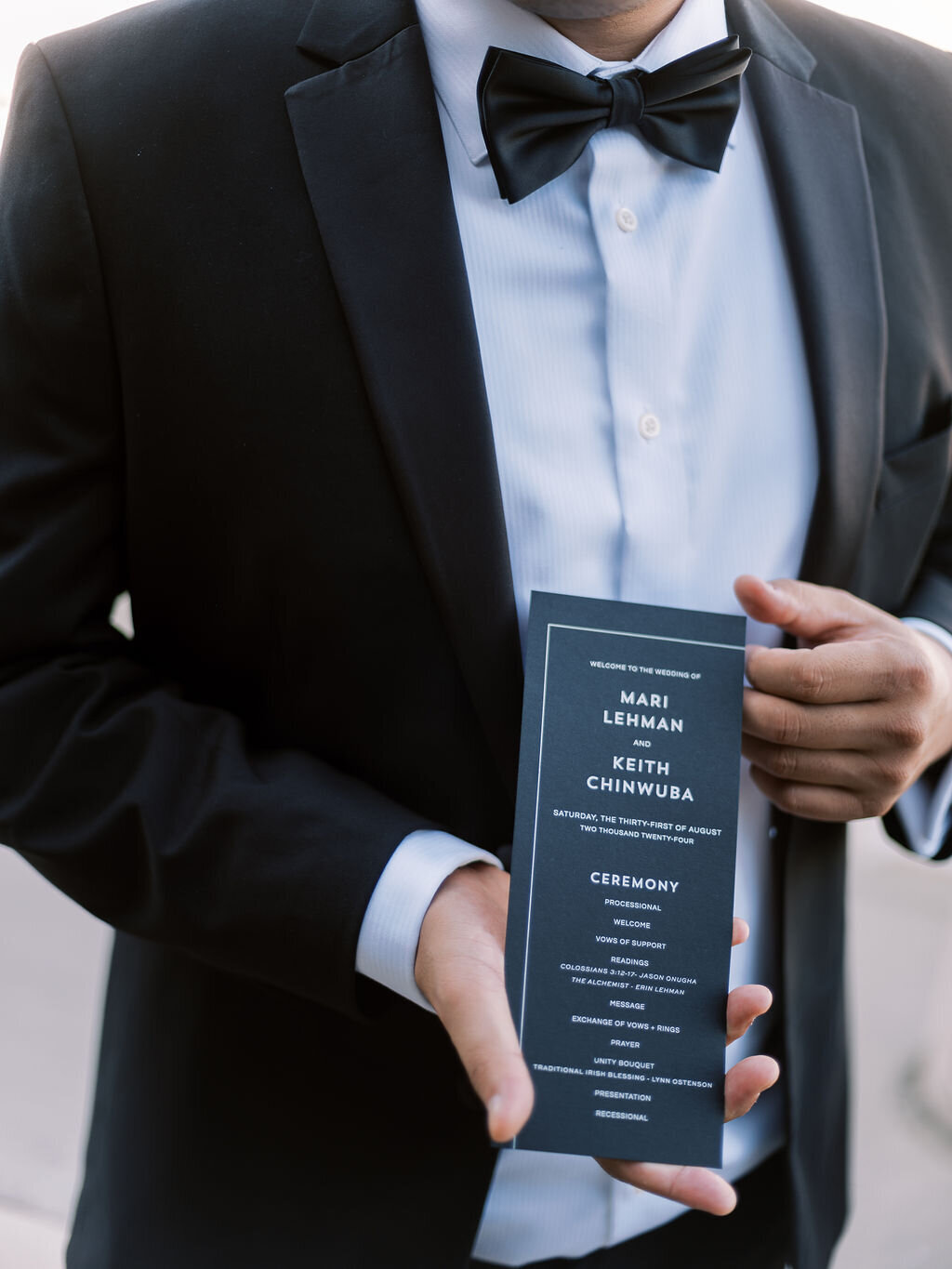 A close-up of a groom holding a sleek black ceremony program featuring the couple’s names and order of events.