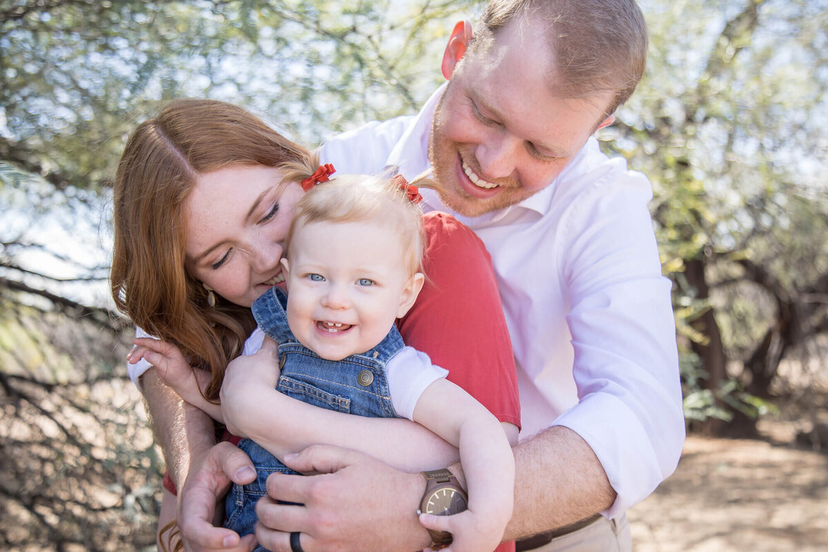 mom and dad snuggling with happy toddler in denim dress