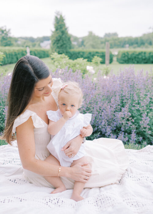 A mom and her one year old daughter sit on a blanket in a field of purple flowers during their Evanston newborn photographer session
