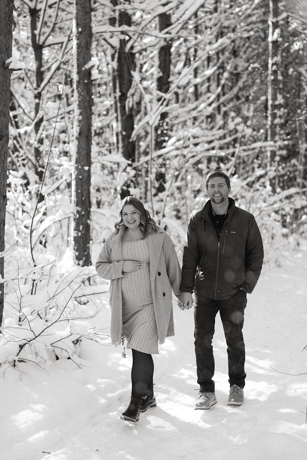 Expecting couple smiling and walking together through the snowy woods at Al Sabo Preserve during their maternity photoshoot.