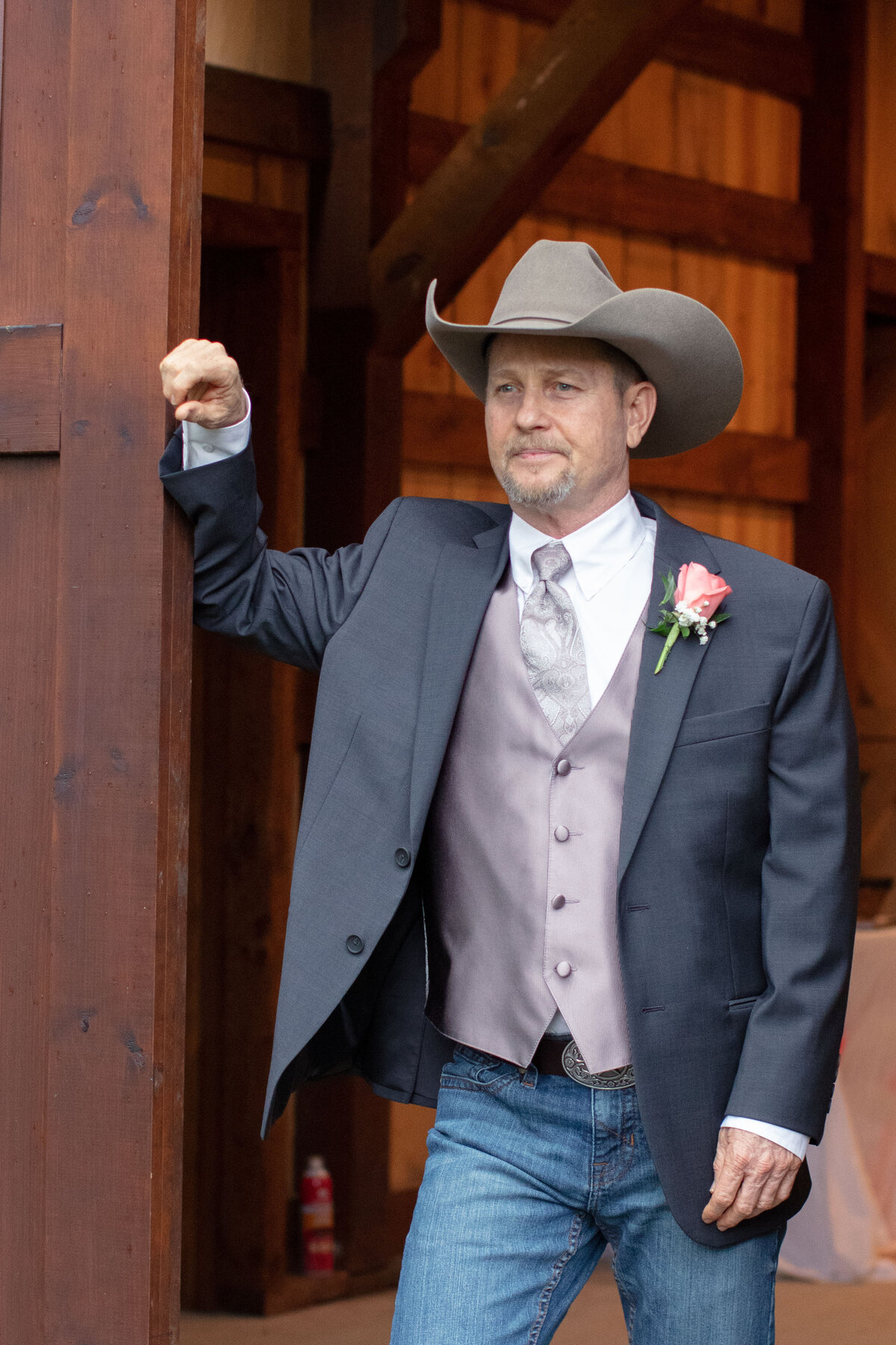 groom in cowboy hat leans against barn doors of rustic wedding in Boerne Texas