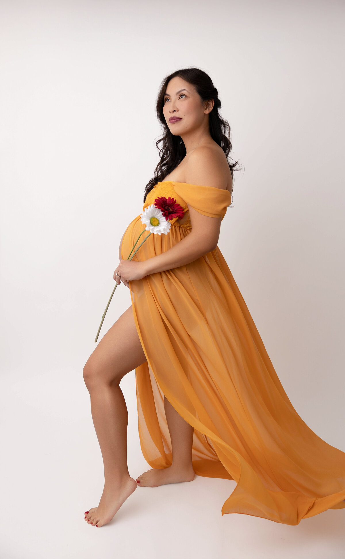 Pregnant mom wearing a yellow off the shoulder dress and posing toward the professional lighting and holding a white and red daisy flower.