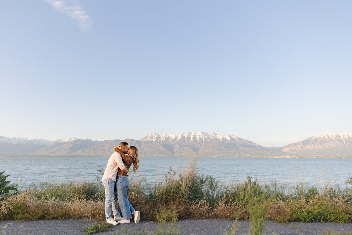 A couple shares a loving embrace near Utah Lake at the Saratoga Springs Marina, with the majestic Mt. Timpanogos in the background. The golden evening light casts a warm glow over the landscape, enhancing the romantic atmosphere.