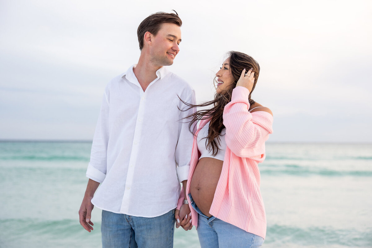 a young married couple with the wife pregnant with their first baby walking on the beach in Destin, FLorida.